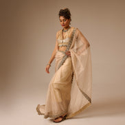 Woman in a traditional beige saree with gold border against a plain background