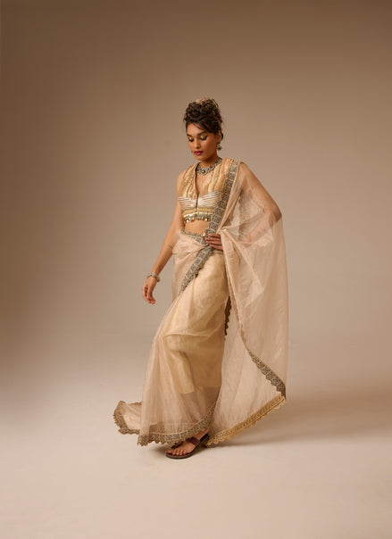 Woman in a traditional beige saree with gold border against a plain background