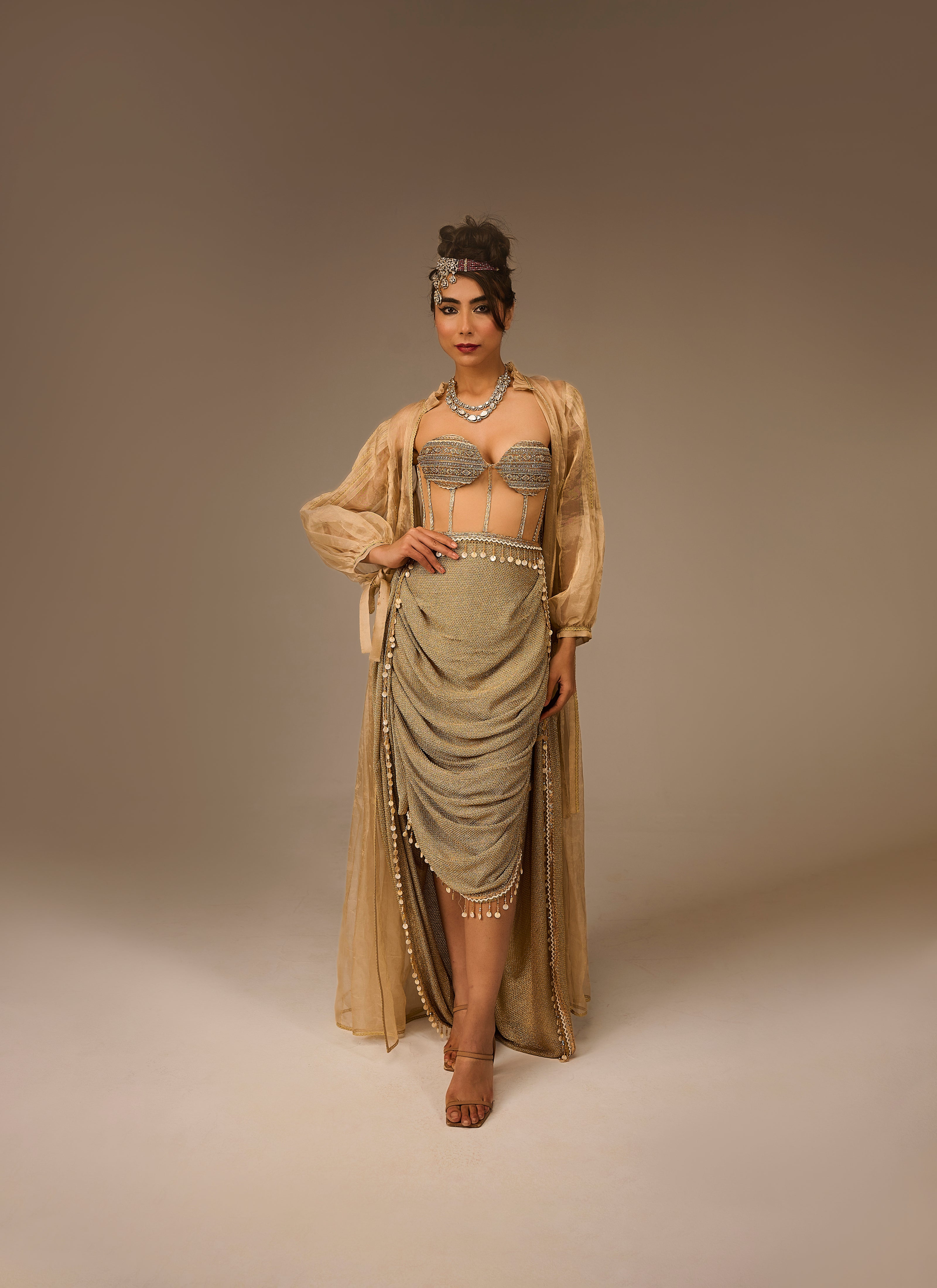 Woman in a flowing beige dress with intricate detailing on a plain background
