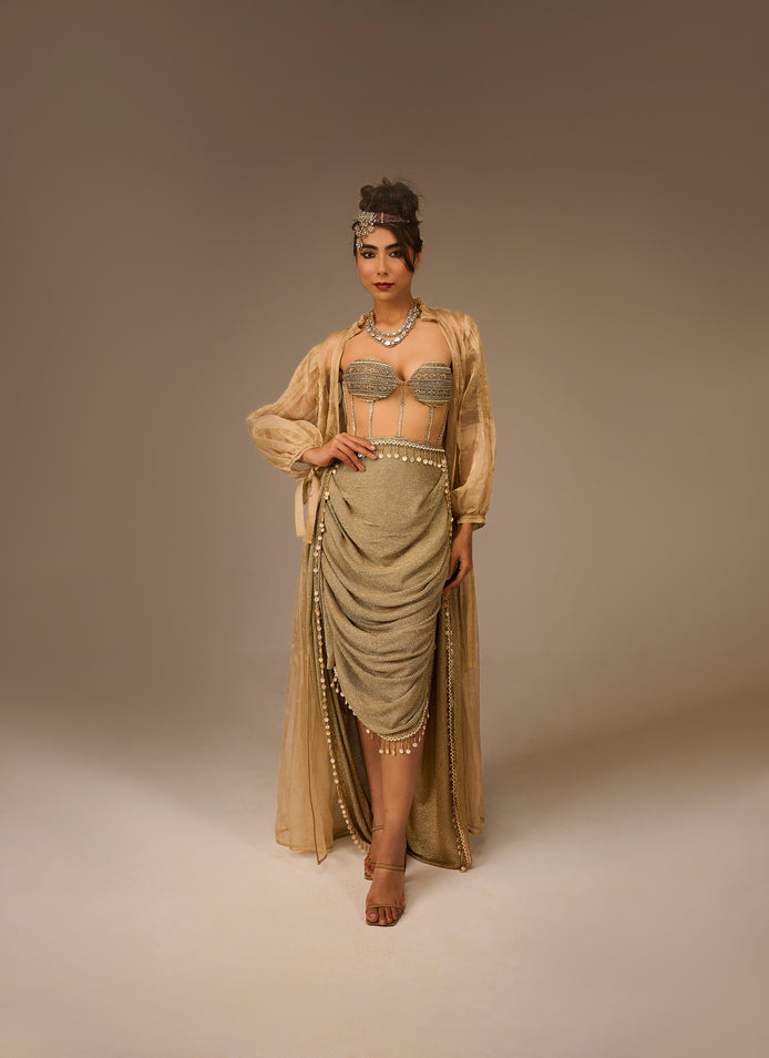 Woman in a flowing beige dress with intricate detailing on a plain background