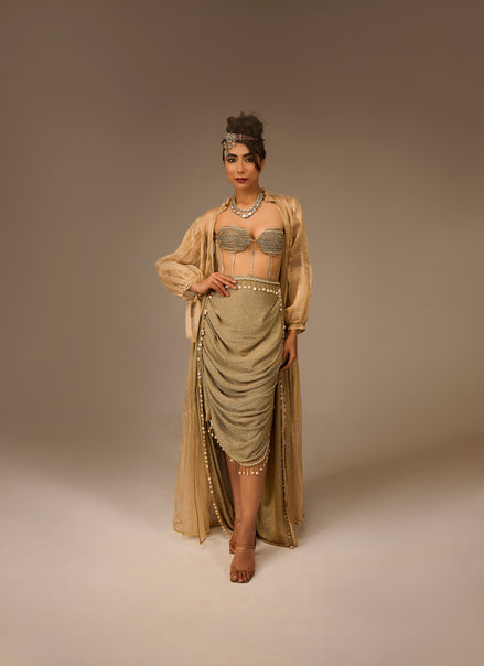 Woman in a flowing beige dress with intricate detailing on a plain background