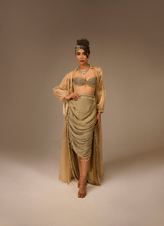 Woman in a flowing beige dress with intricate detailing on a plain background