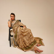 Woman in a traditional embroidered saree sitting on a chair against a plain background