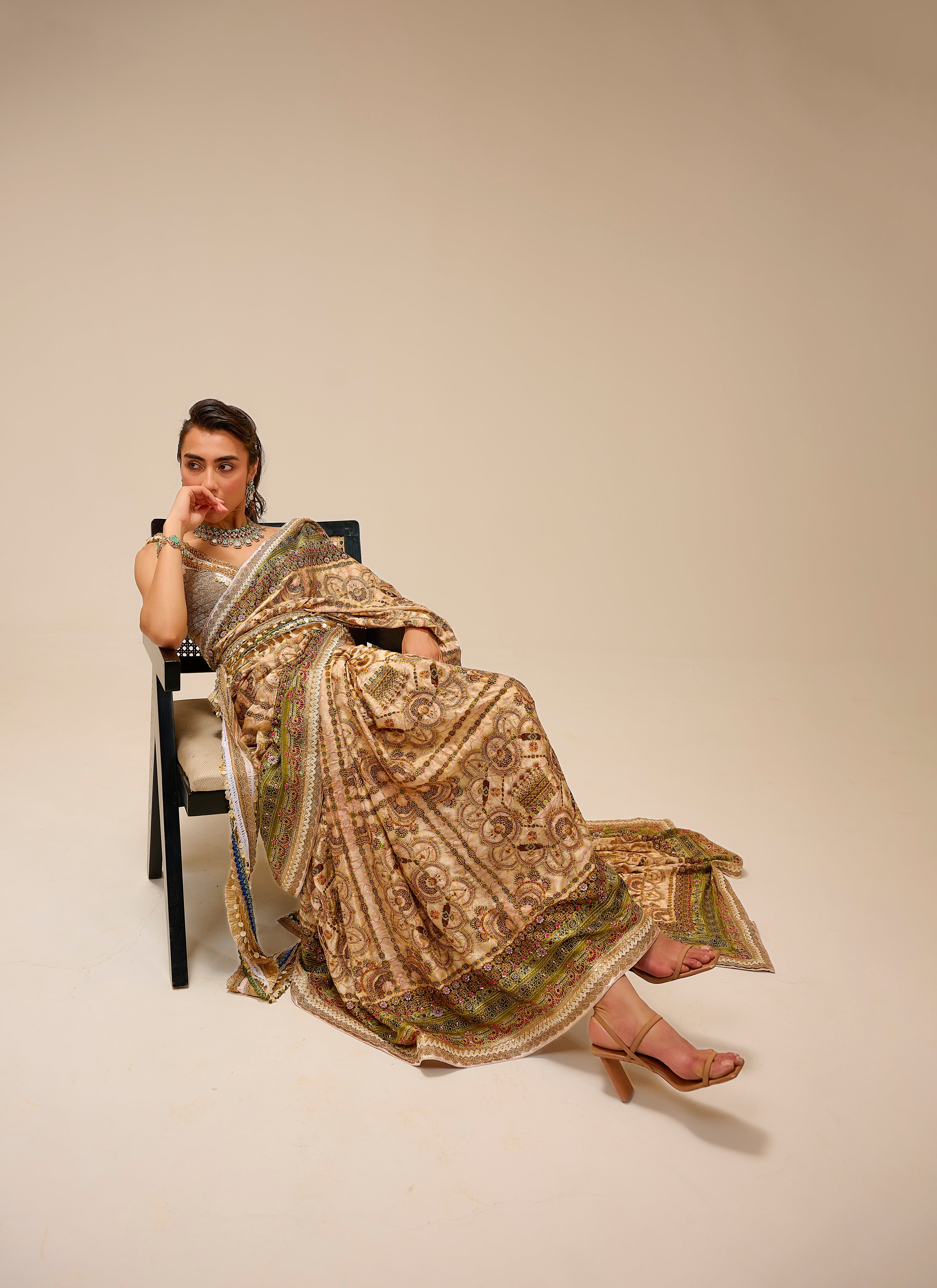 Woman in a traditional embroidered saree sitting on a chair against a plain background