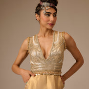 Woman wearing a gold beaded top and beige pants against a brown background