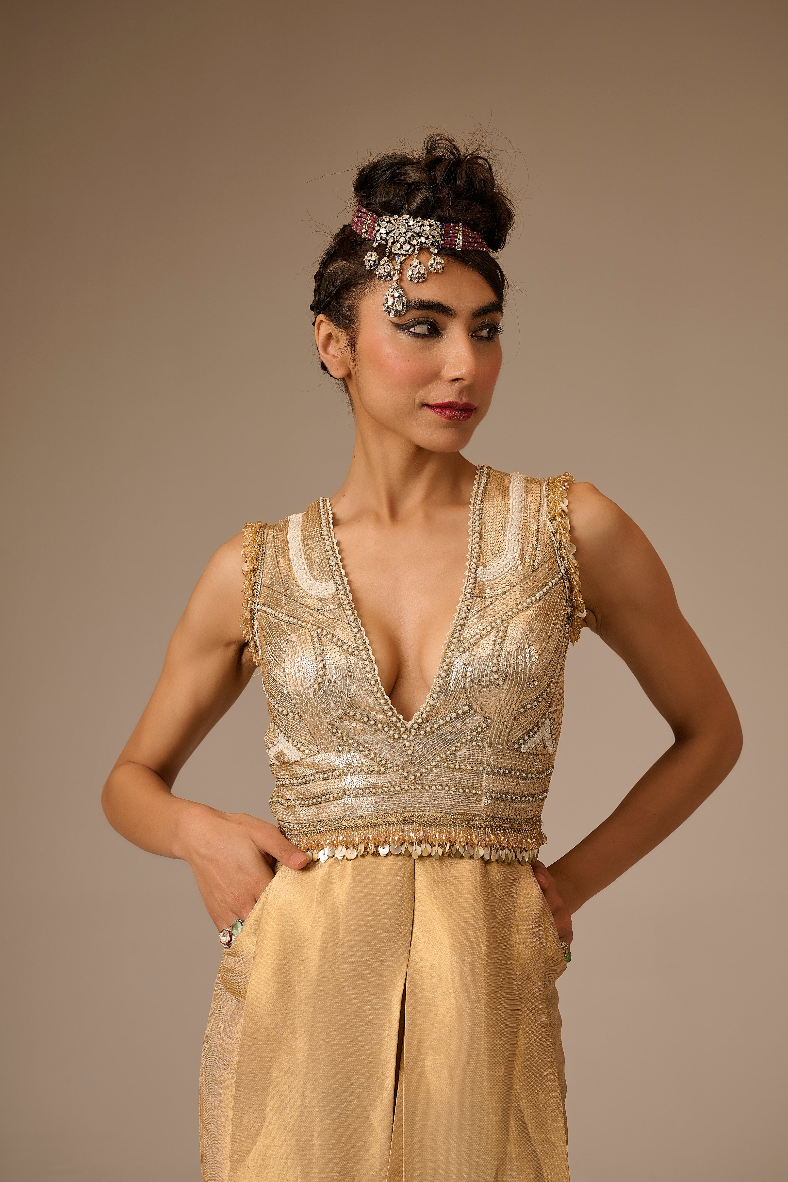 Woman wearing a gold beaded top and beige pants against a brown background