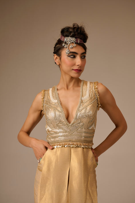 Woman wearing a gold beaded top and beige pants against a brown background