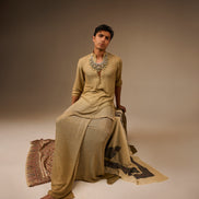 Person wearing a traditional outfit with jewelry, sitting on a patterned rug against a plain background