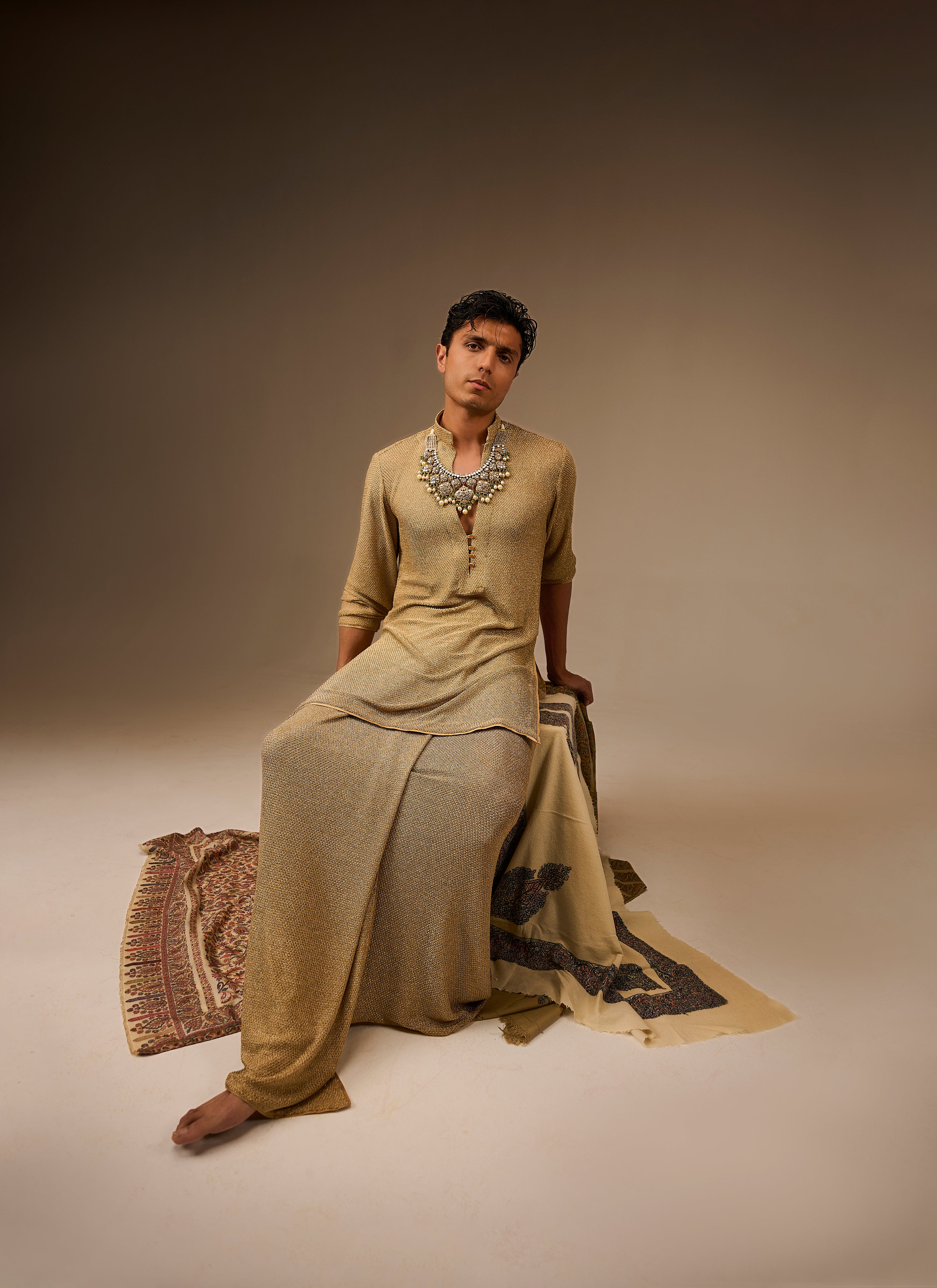 Person wearing a traditional outfit with jewelry, sitting on a patterned rug against a plain background