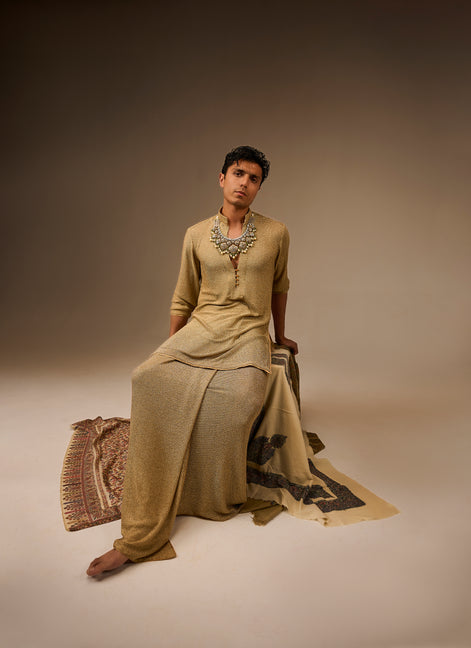 Person wearing a traditional outfit with jewelry, sitting on a patterned rug against a plain background