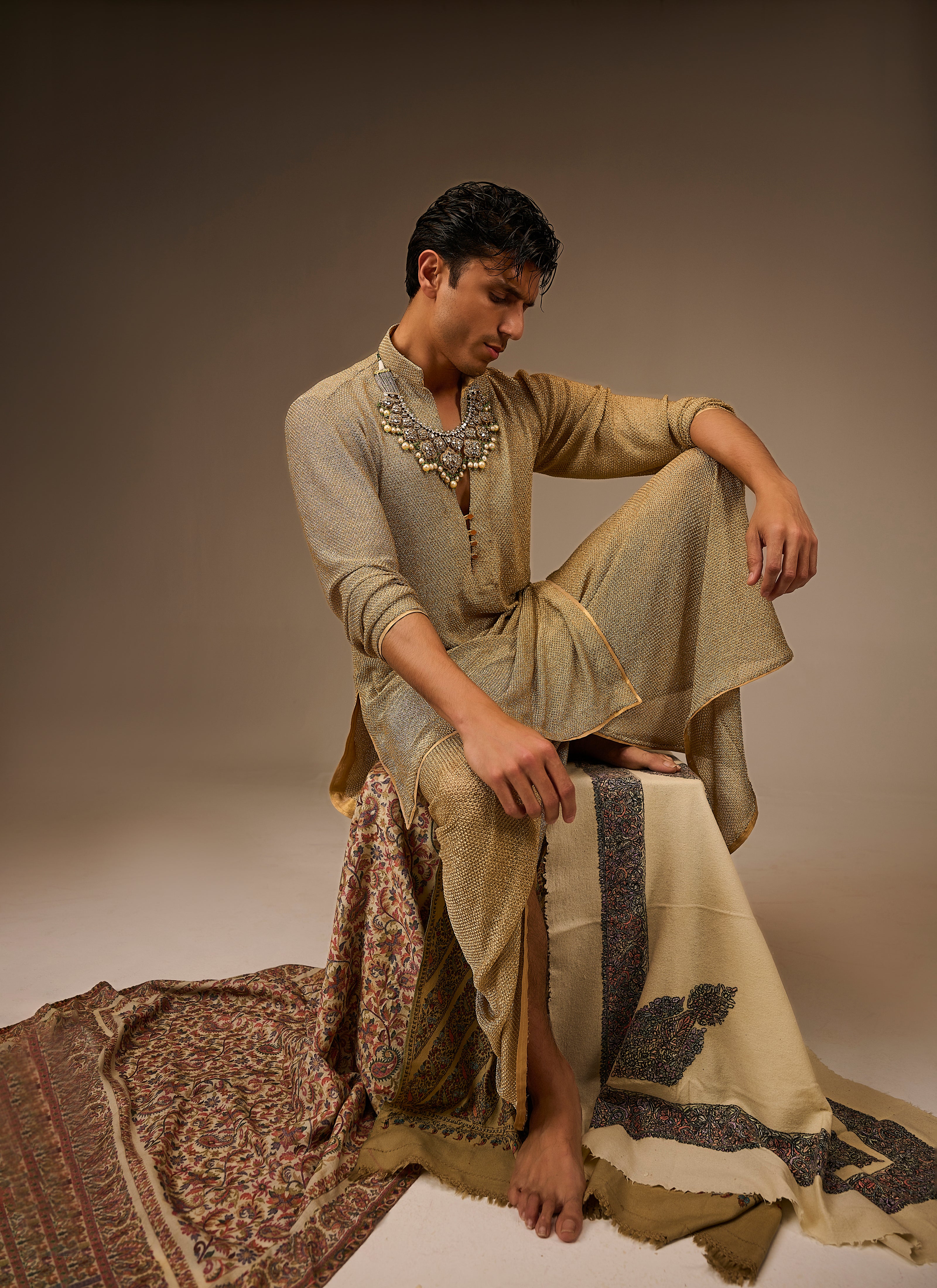 Man in traditional attire sitting on a patterned rug against a plain background