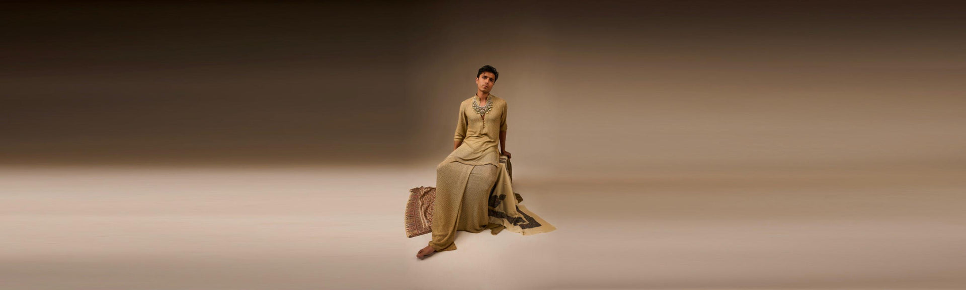 Man in a long, light-colored dress sitting on a beige platform with a plain background
